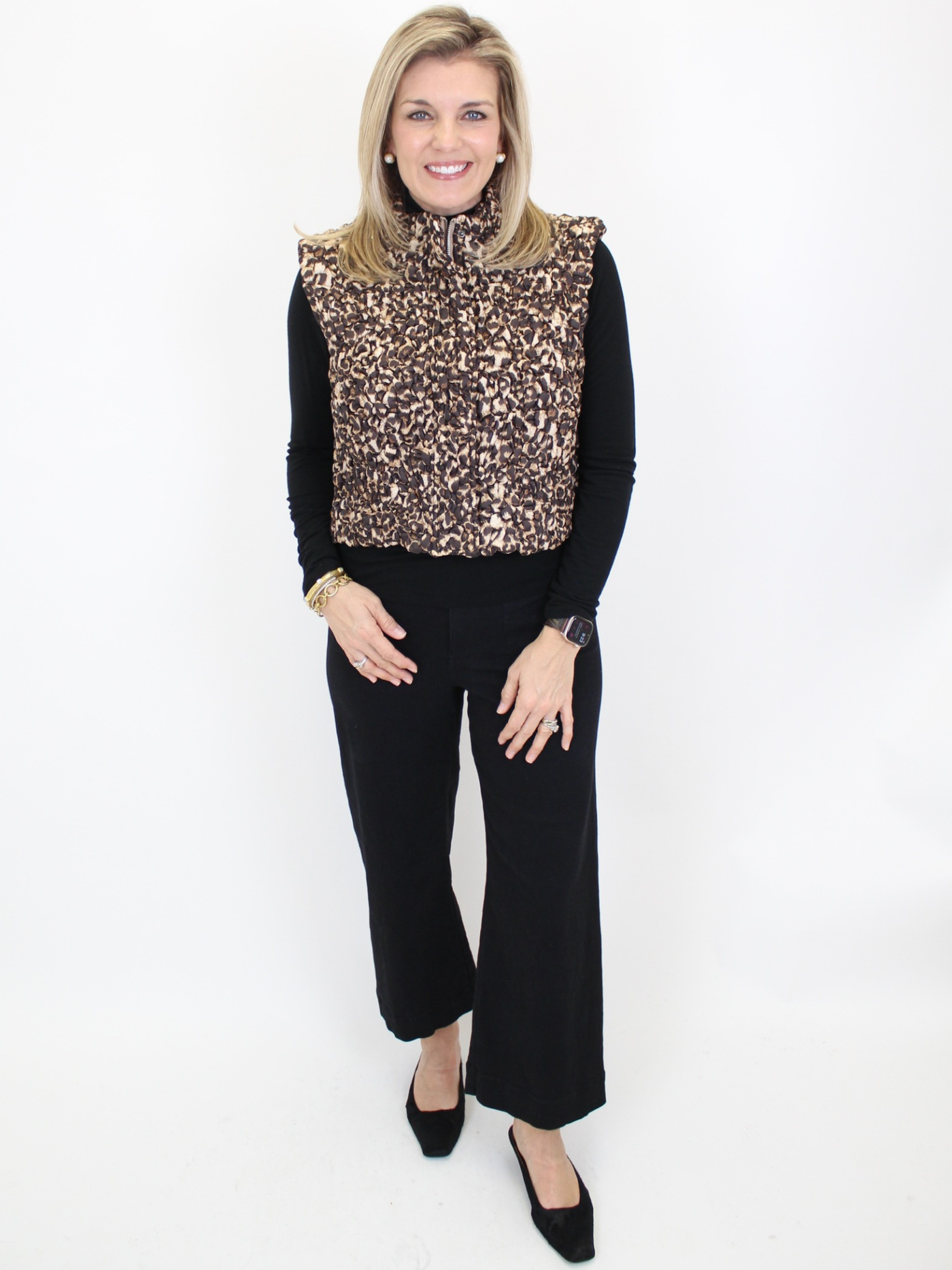Woman wearing a leopard print vest over a black long-sleeve shirt and black pants on a white background