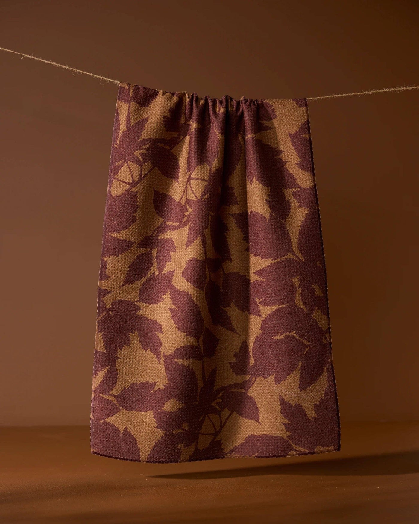 Brown leaf print tea towel hanging against a brown background