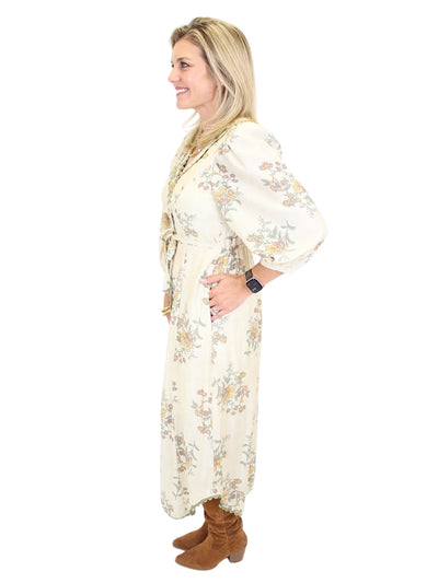 Woman wearing a long cream floral dress on a white background, side.