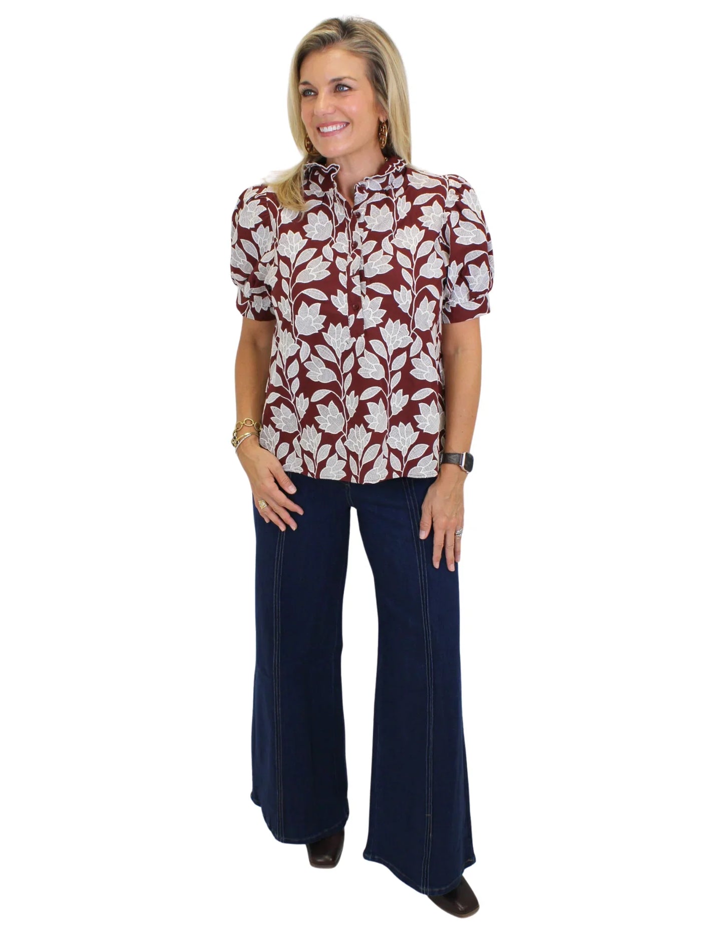 Model wearing burgundy floral top with flared jeans on white background, side.