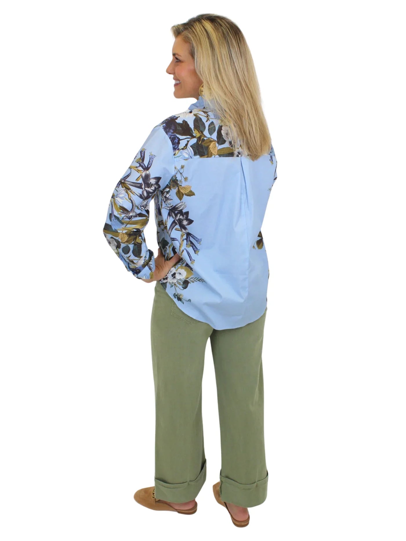 Model wearing blue botanical print button down top with green pants on white background, back.