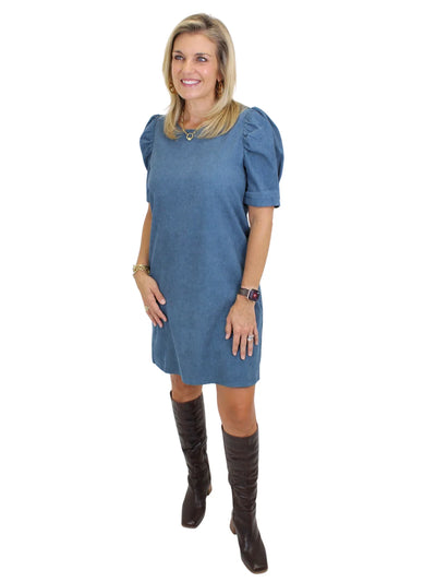 Model wearing a blue dress with puffed sleeves and brown knee-high boots on a white background, side.