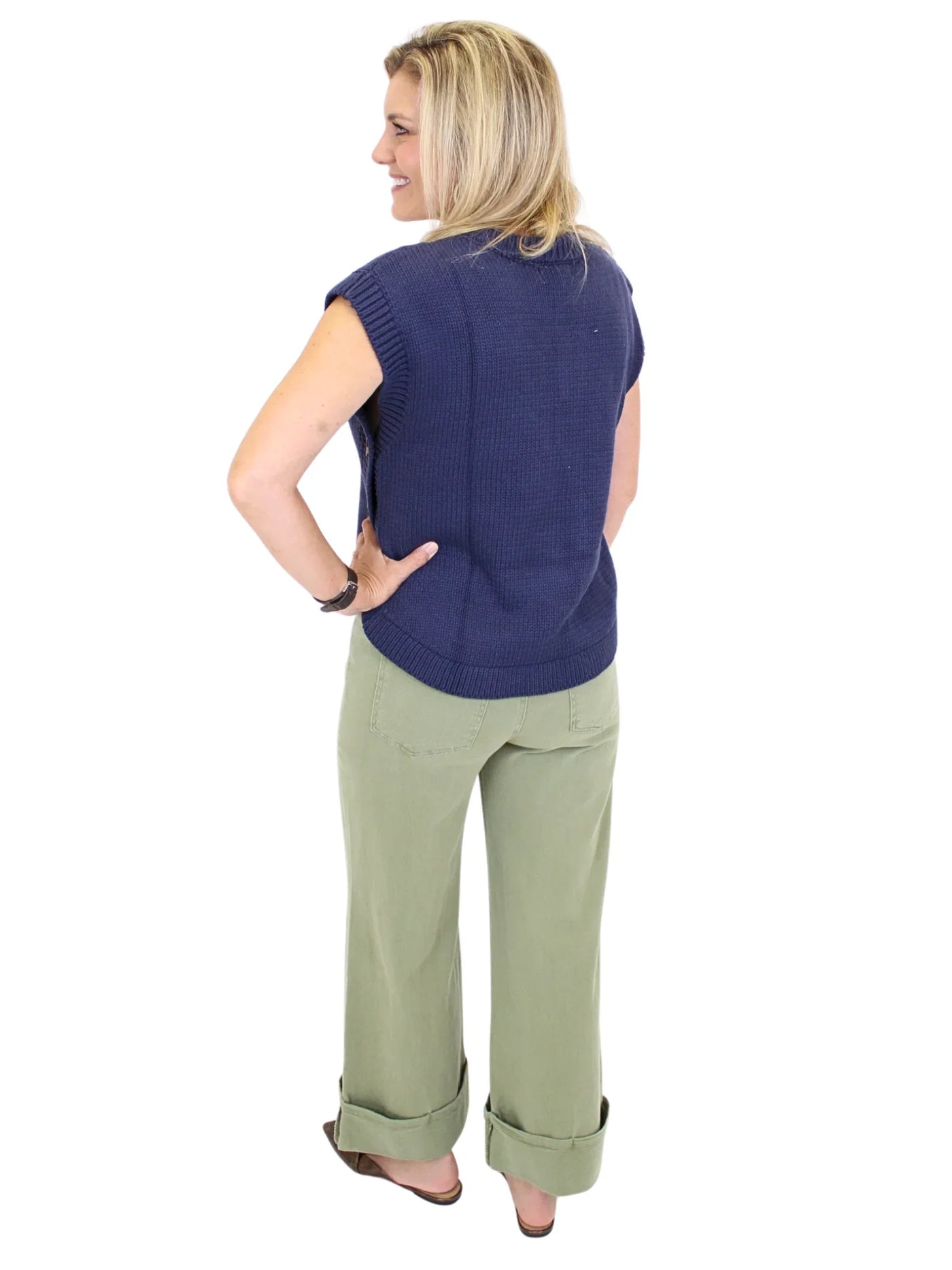 Woman wearing a navy blue sleeveless sweater and green pants on a white background, back.