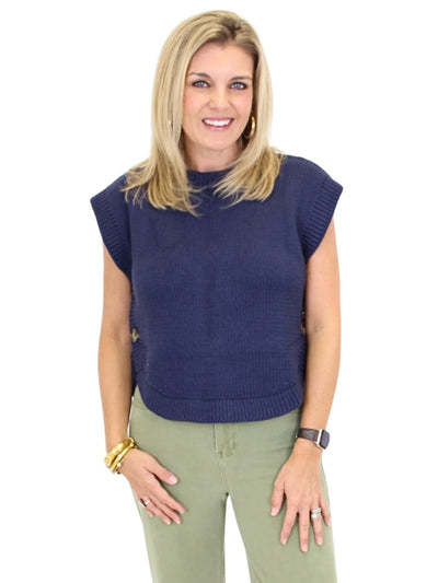 Woman wearing a navy blue sleeveless sweater and green pants on a white background, closeup front.