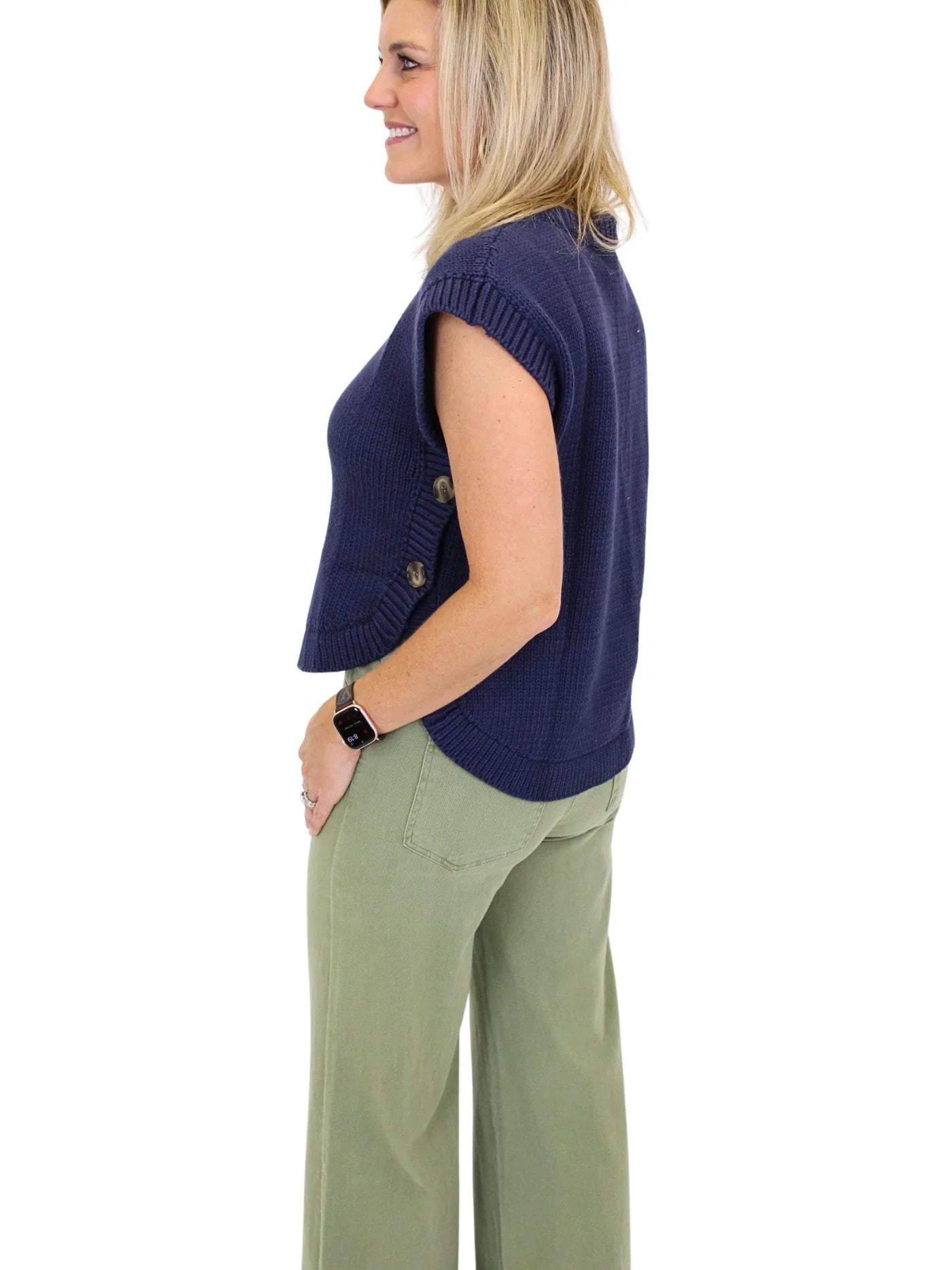 Woman wearing a navy blue sleeveless sweater and green pants on a white background, side.