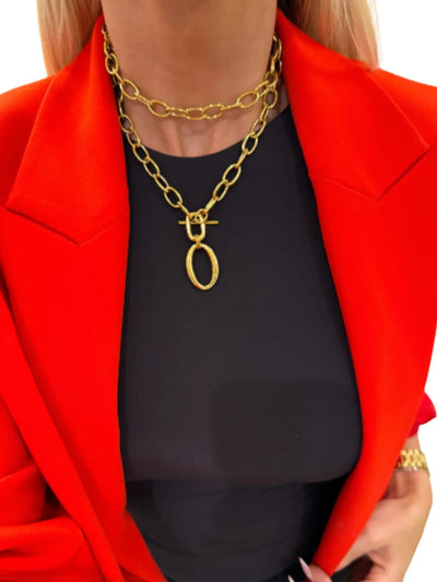 Model in red blazer wearing layered gold chain necklace with toggle and oval pendant.