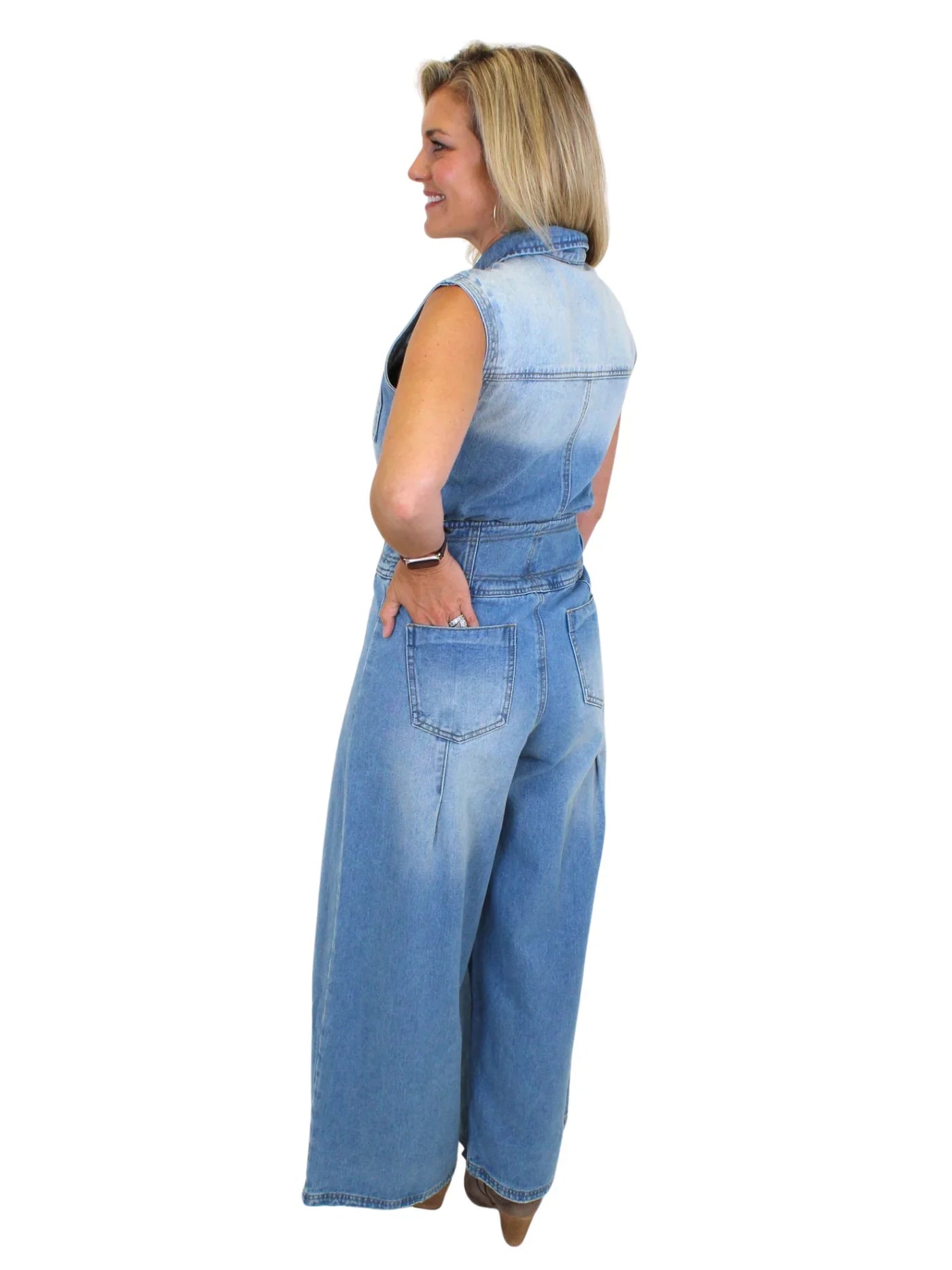 Model wearing wide leg denim jumpsuit with hand in back pocket on white background, back.