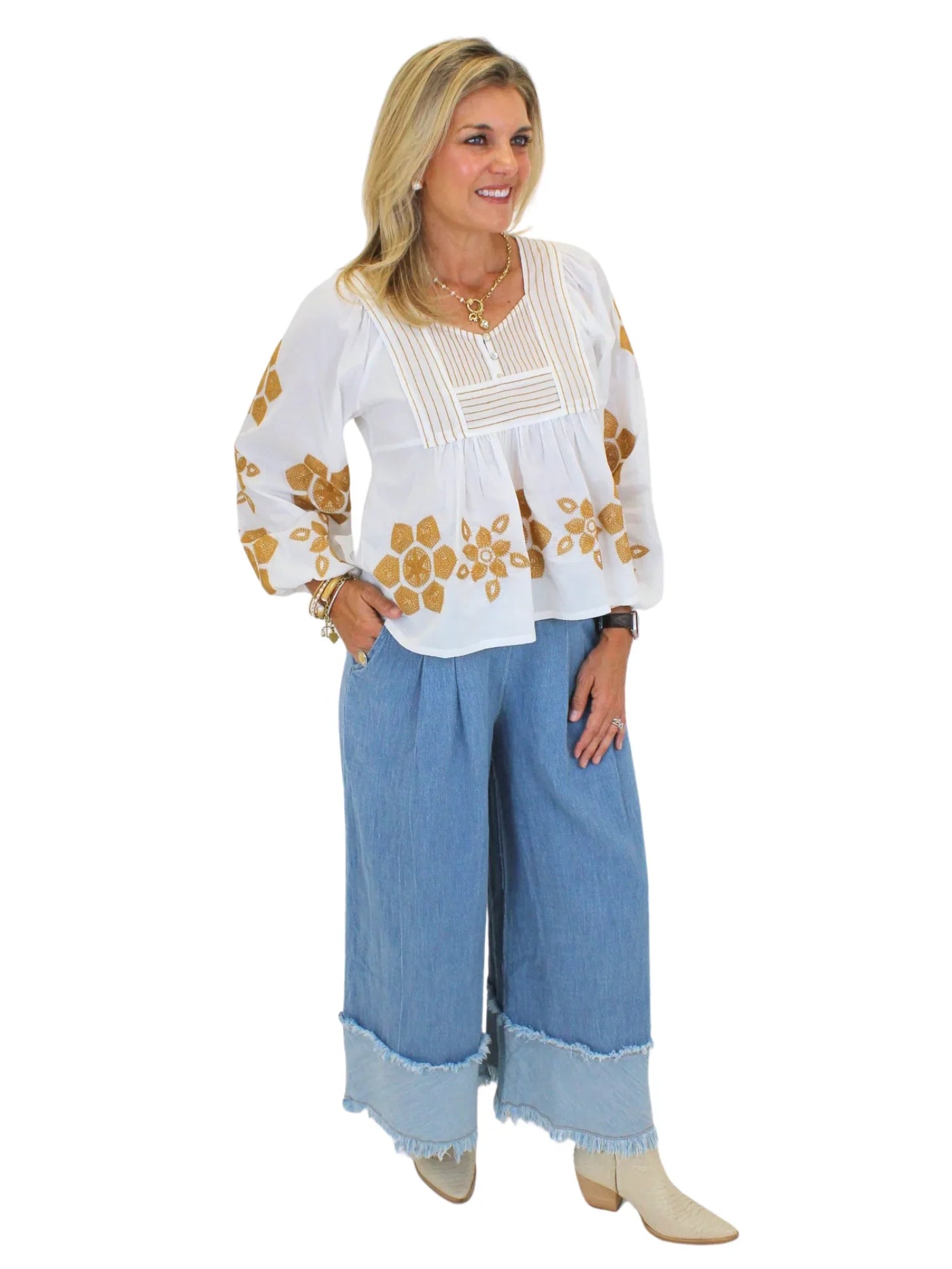 Model wearing tan embroidered white babydoll blouse and wide leg blue jeans on white background, side.